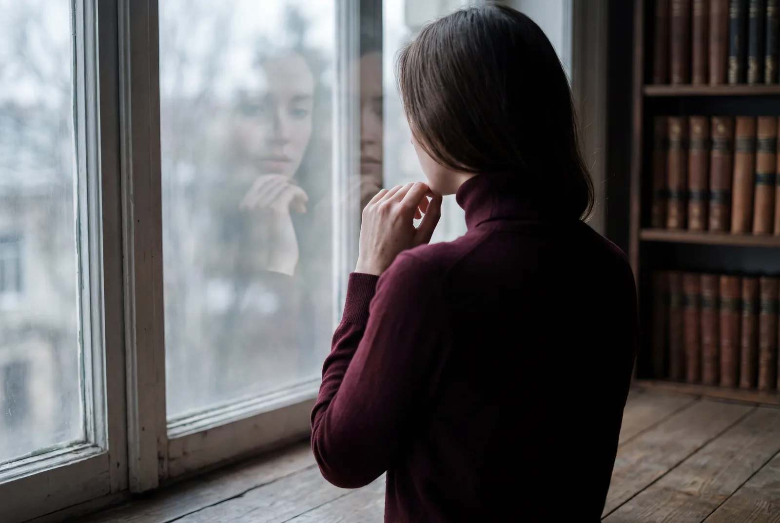 Femme de dos face à une fenêtre, son reflet dédoublé dans la vitre